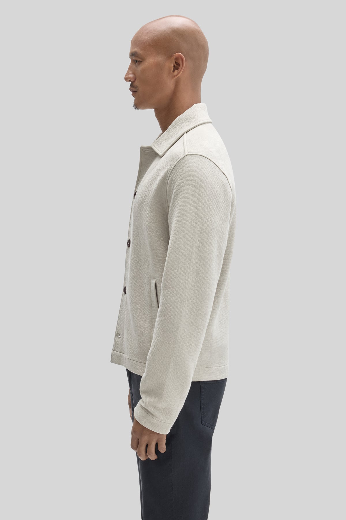 Cream-colored buttoned jacket with a collar, worn over dark trousers, shown from the side.