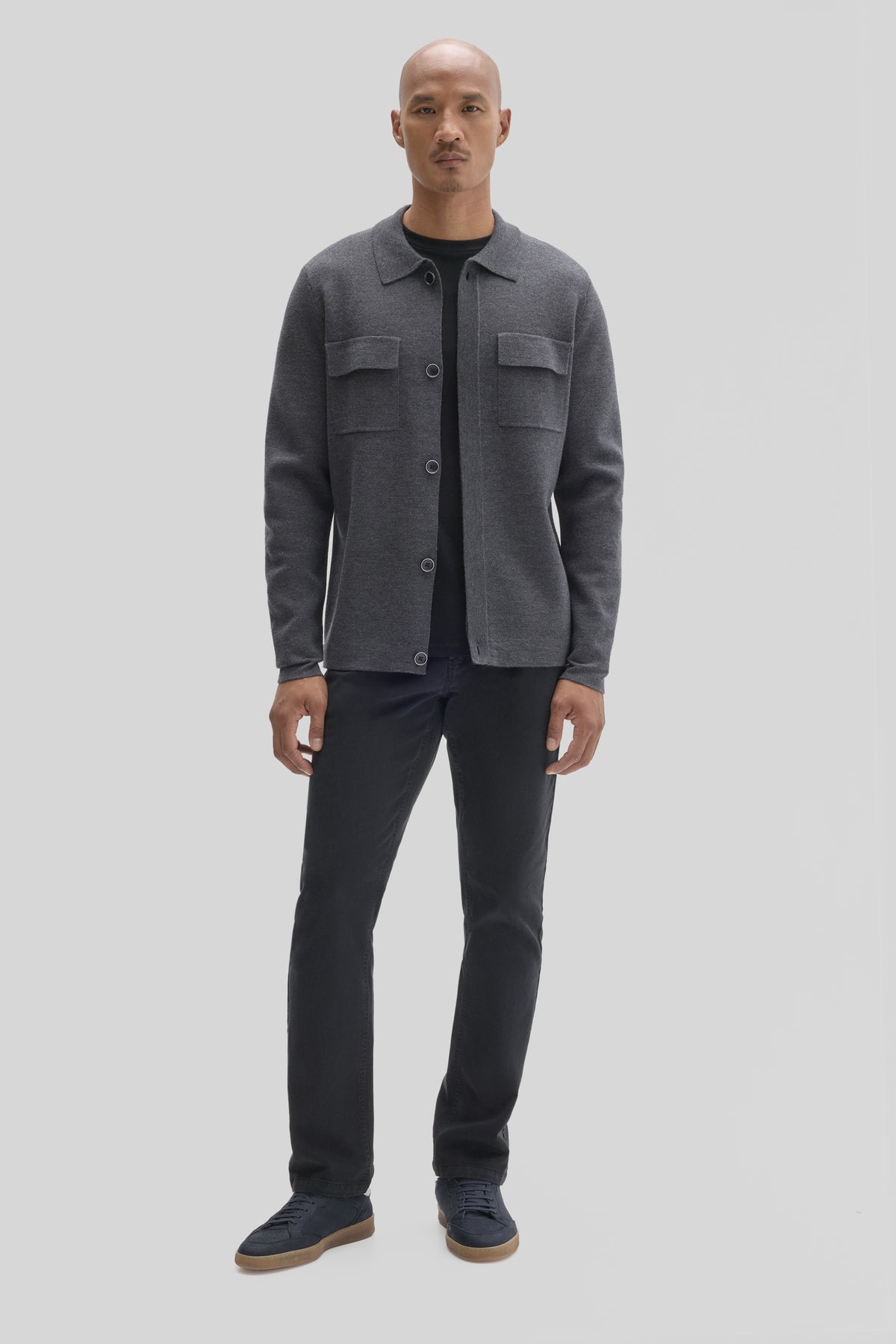Man wearing charcoal gray buttoned cardigan with chest pockets over black shirt, paired with black jeans and navy sneakers.