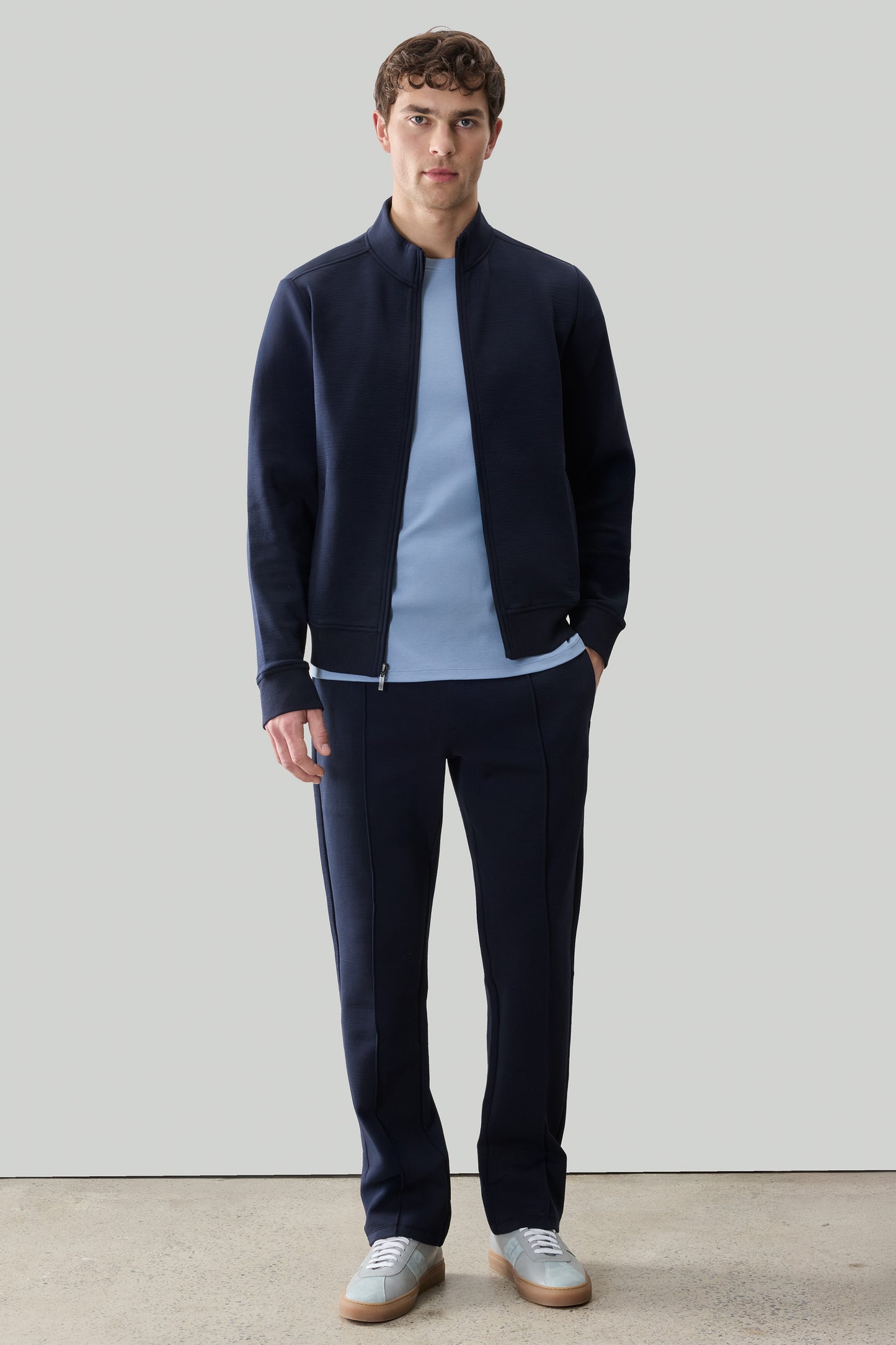 Man wearing navy full-zip long sleeve jacket over light blue t-shirt and navy pants with white sneakers.