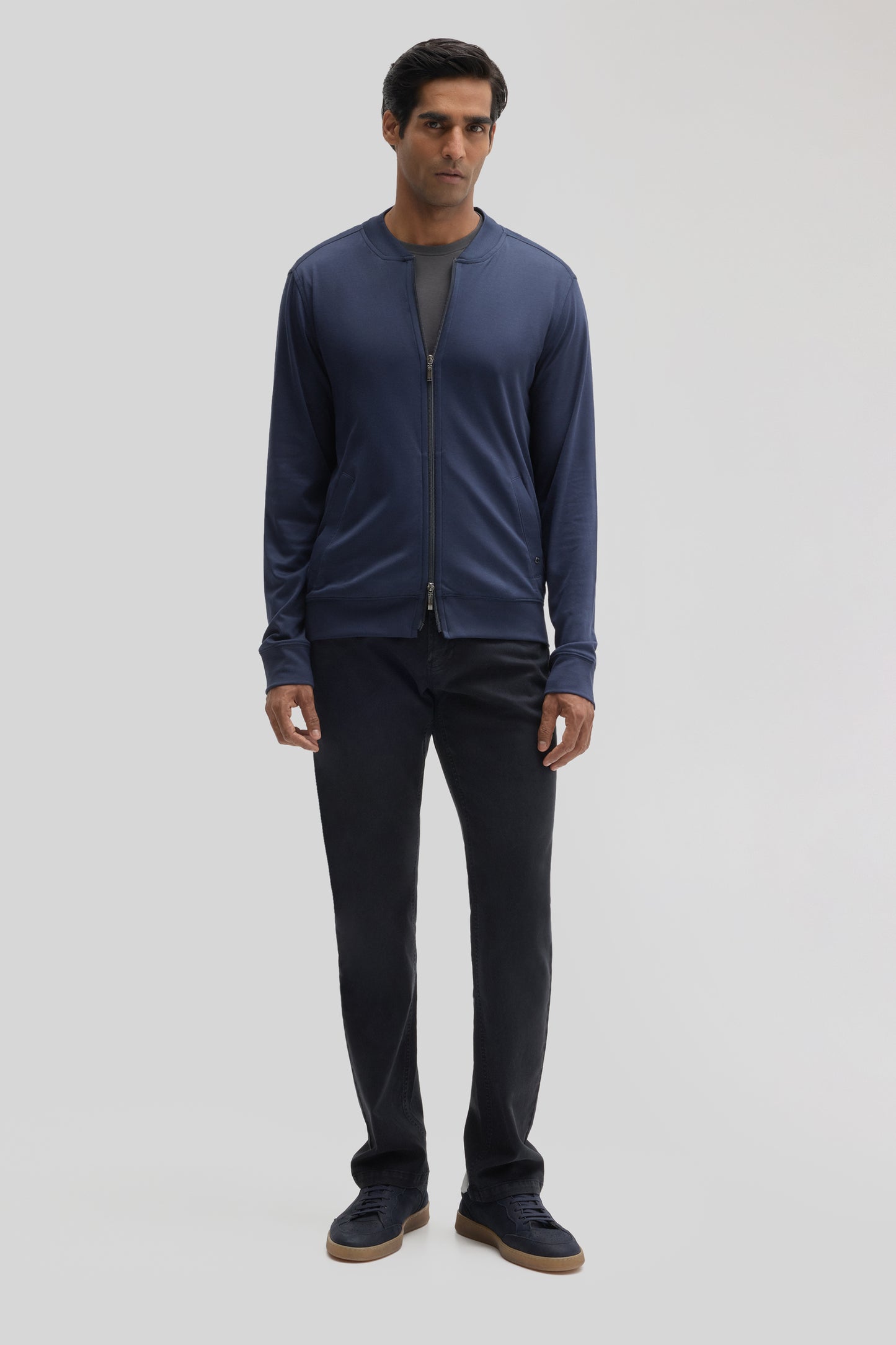 Man wearing navy zip-up baseball jacket over gray shirt, paired with black pants and navy sneakers.