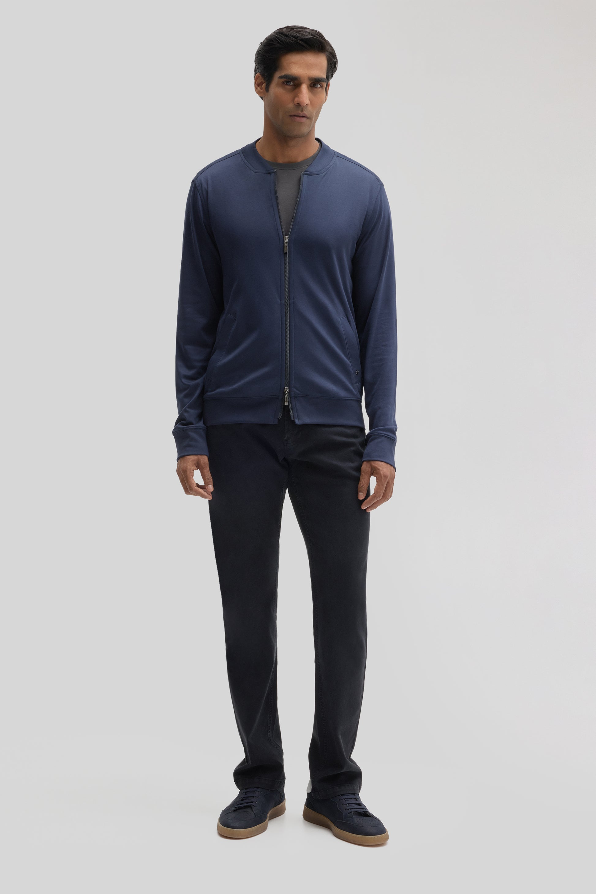 Man wearing navy zip-up baseball jacket over gray shirt, paired with black pants and navy sneakers.