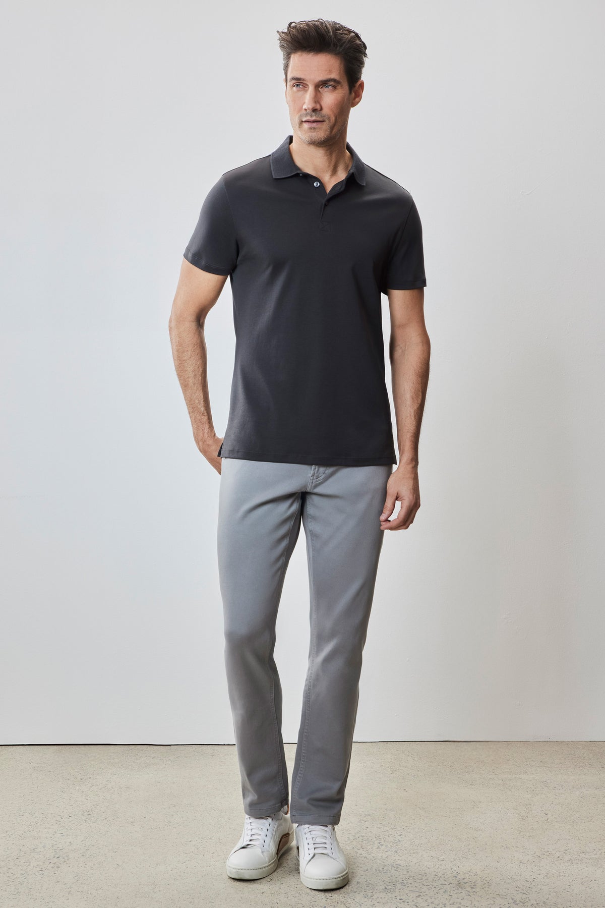 Man wearing black short-sleeve polo shirt with white button placket, gray pants, and white sneakers.