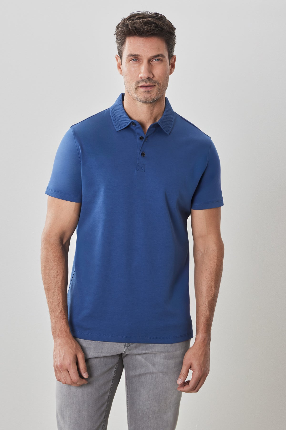 Man wearing a blue short-sleeve polo shirt with button placket and gray trousers against a light gray background.