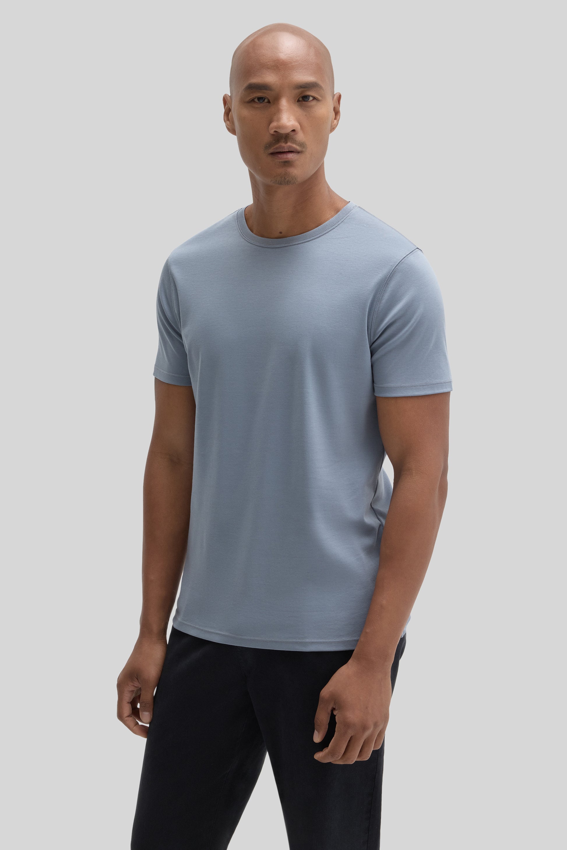 Man wearing a light blue crew neck t-shirt with short sleeves and black pants against a gray background.