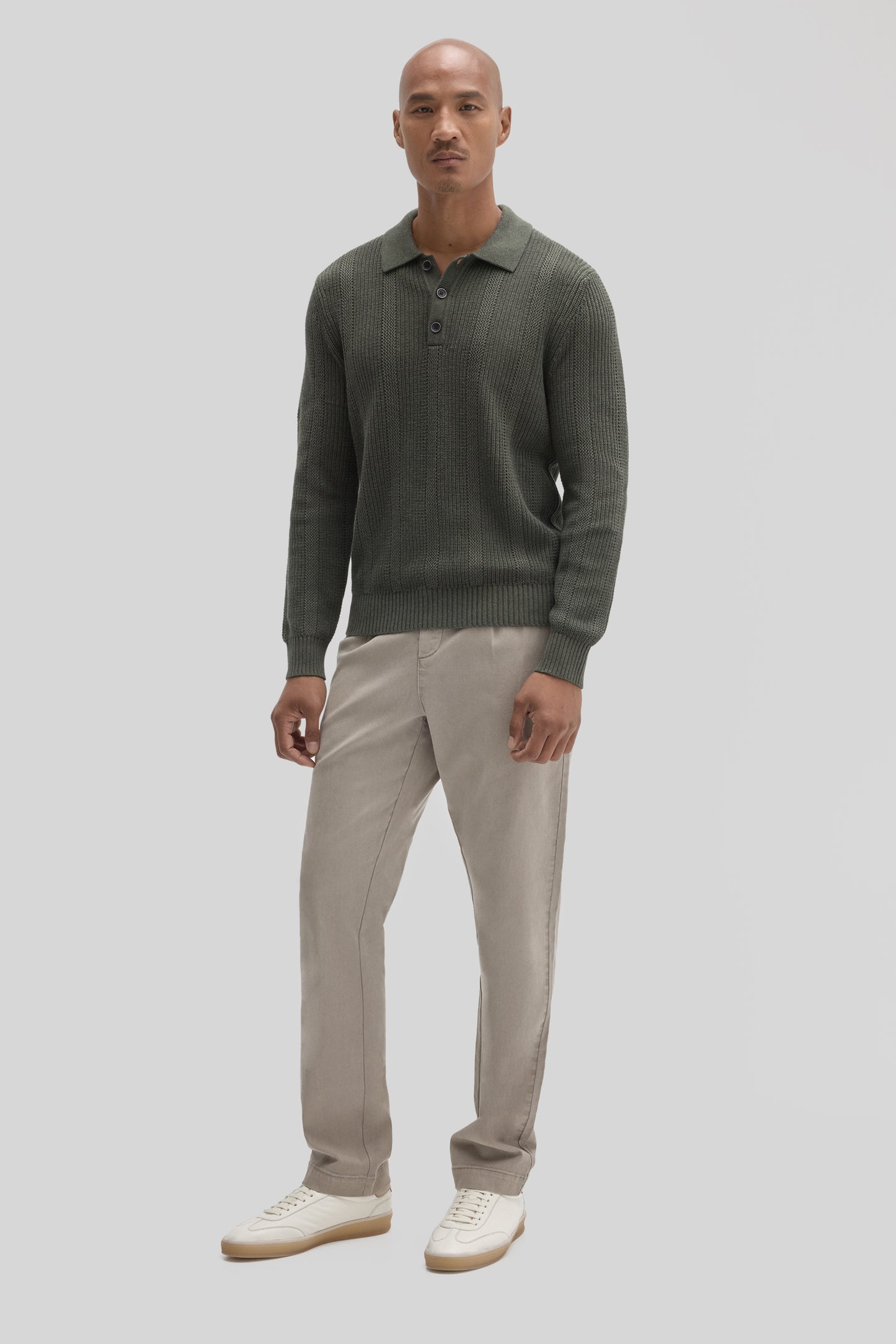 Man wearing olive green ribbed sweater polo with collar and buttons, paired with beige pants and white sneakers.