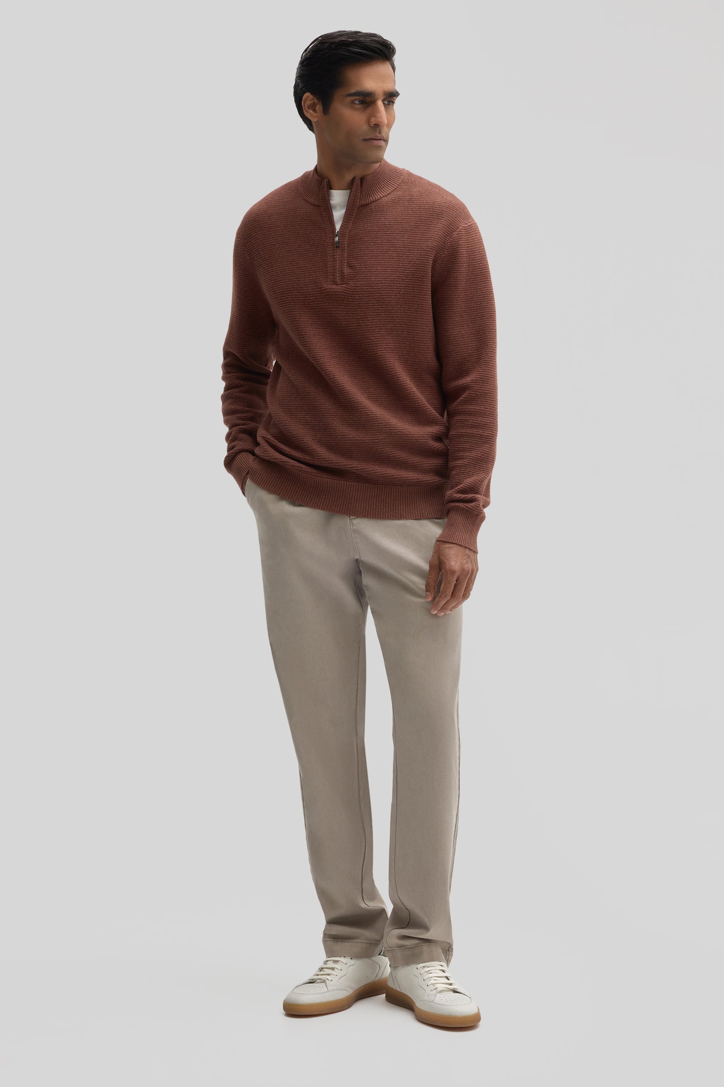 Man wearing rust-colored quarter zip sweater with ribbed texture, cream pants, and white sneakers.