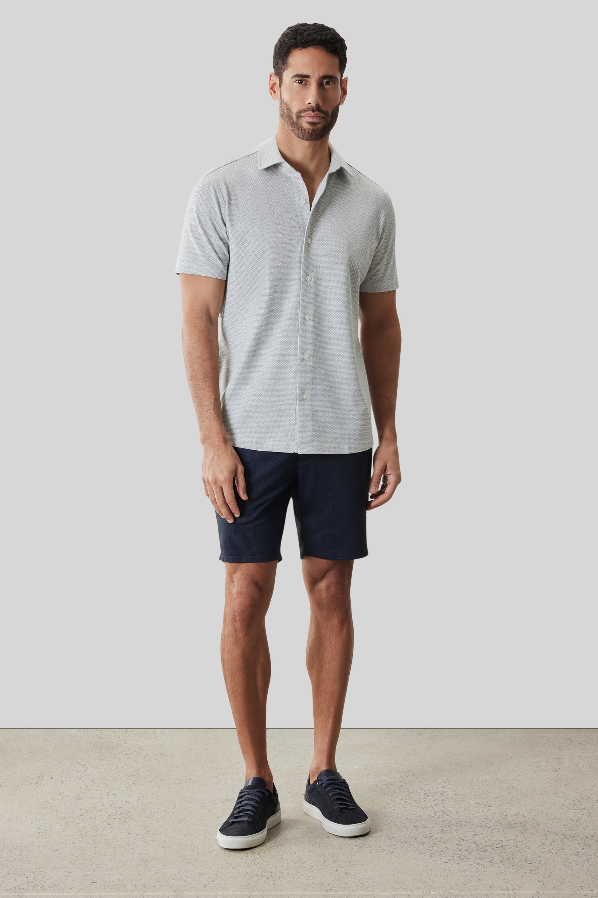 Man wearing light gray short-sleeve button-up knit shirt with navy shorts and dark sneakers.