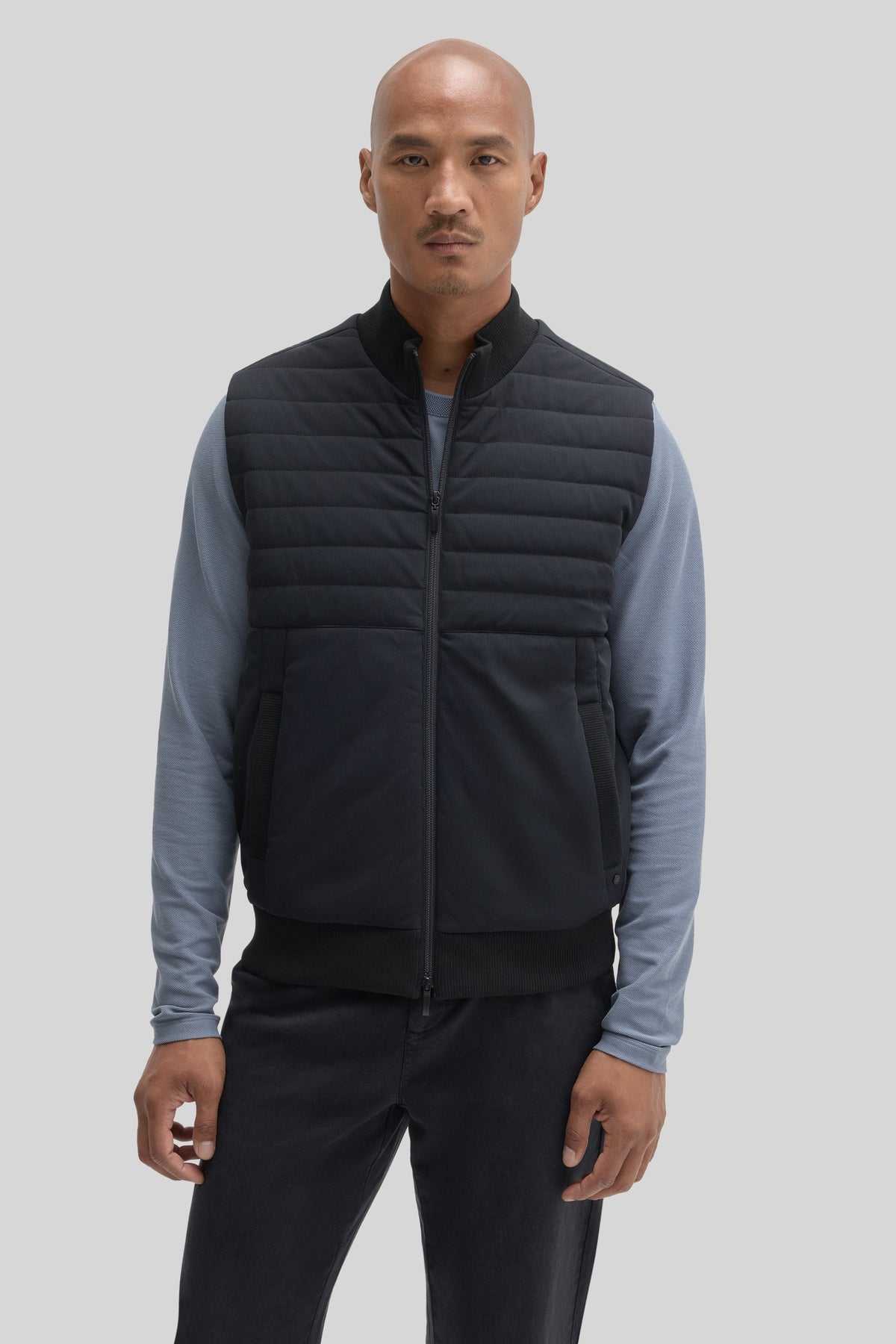 Man wearing black quilted vest with horizontal stitching over blue-gray long-sleeve shirt and dark pants.