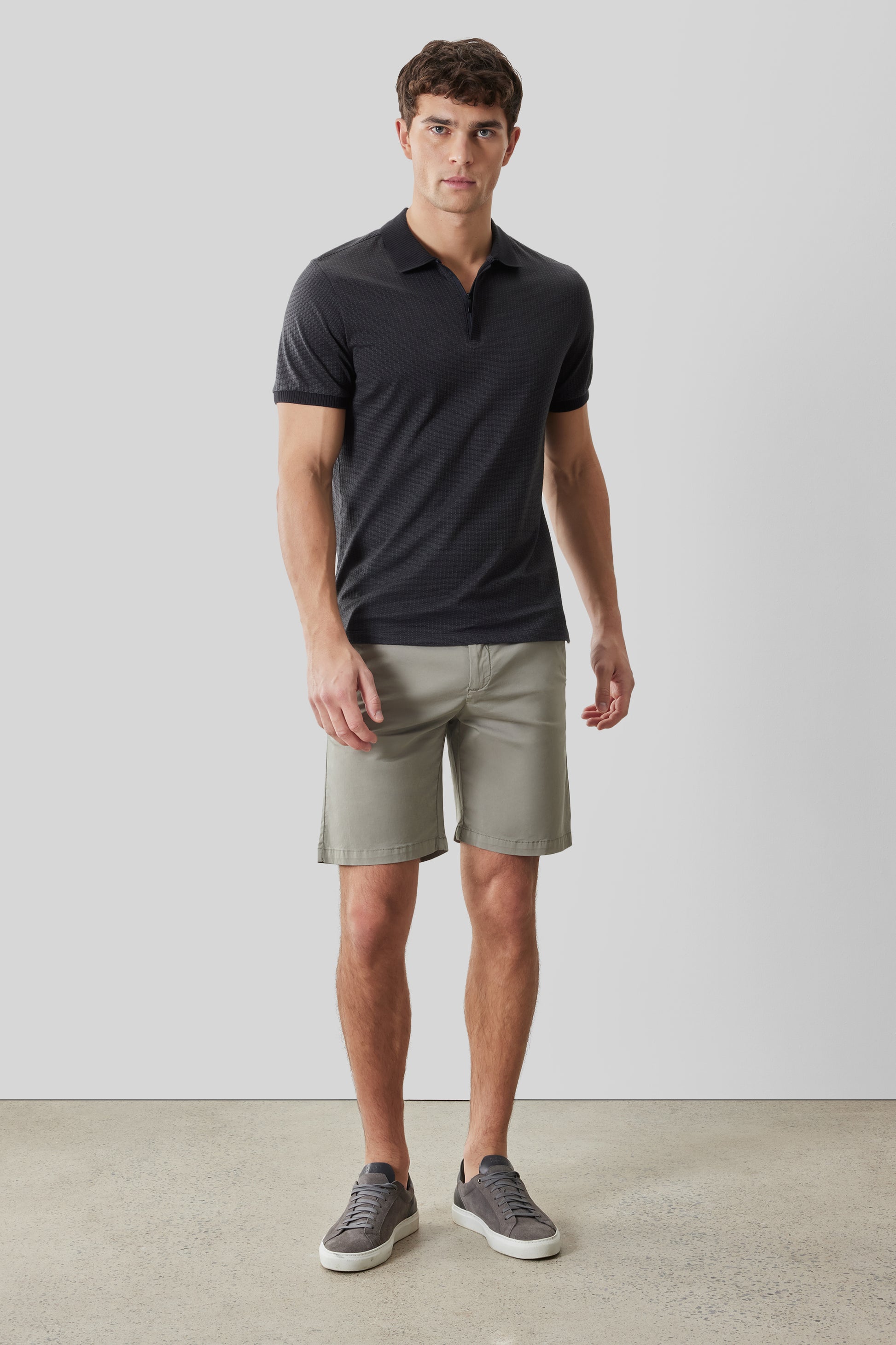 Man wearing dark charcoal short-sleeve polo with covered placket and beige shorts, styled with gray sneakers.