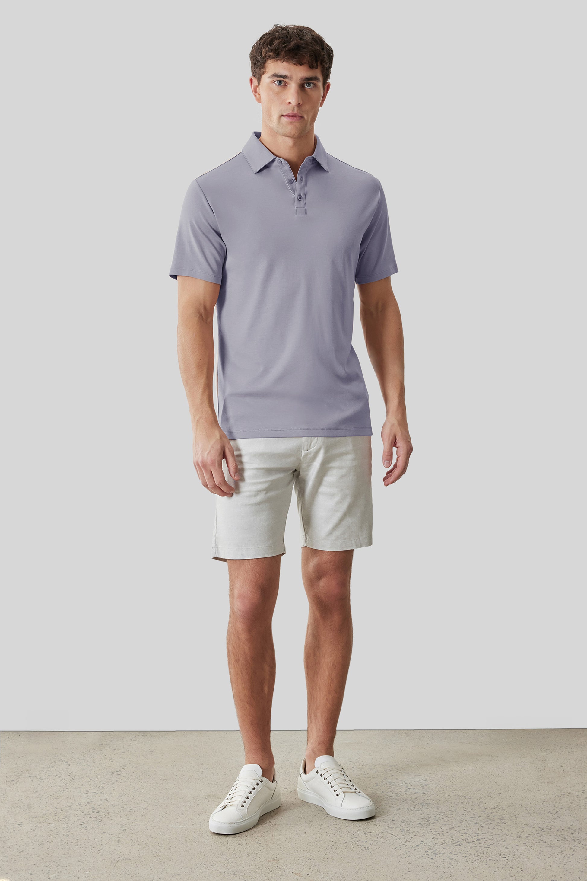 Man wearing lavender herringbone polo shirt with button collar and cream shorts with white sneakers.