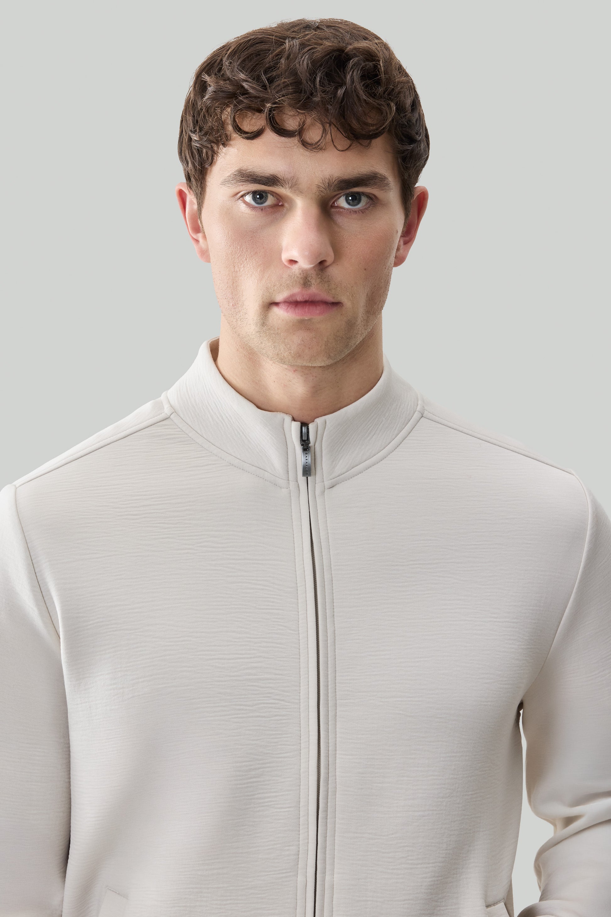 Man wearing cream-colored long sleeve full zip jacket with textured fabric and mock neck collar.