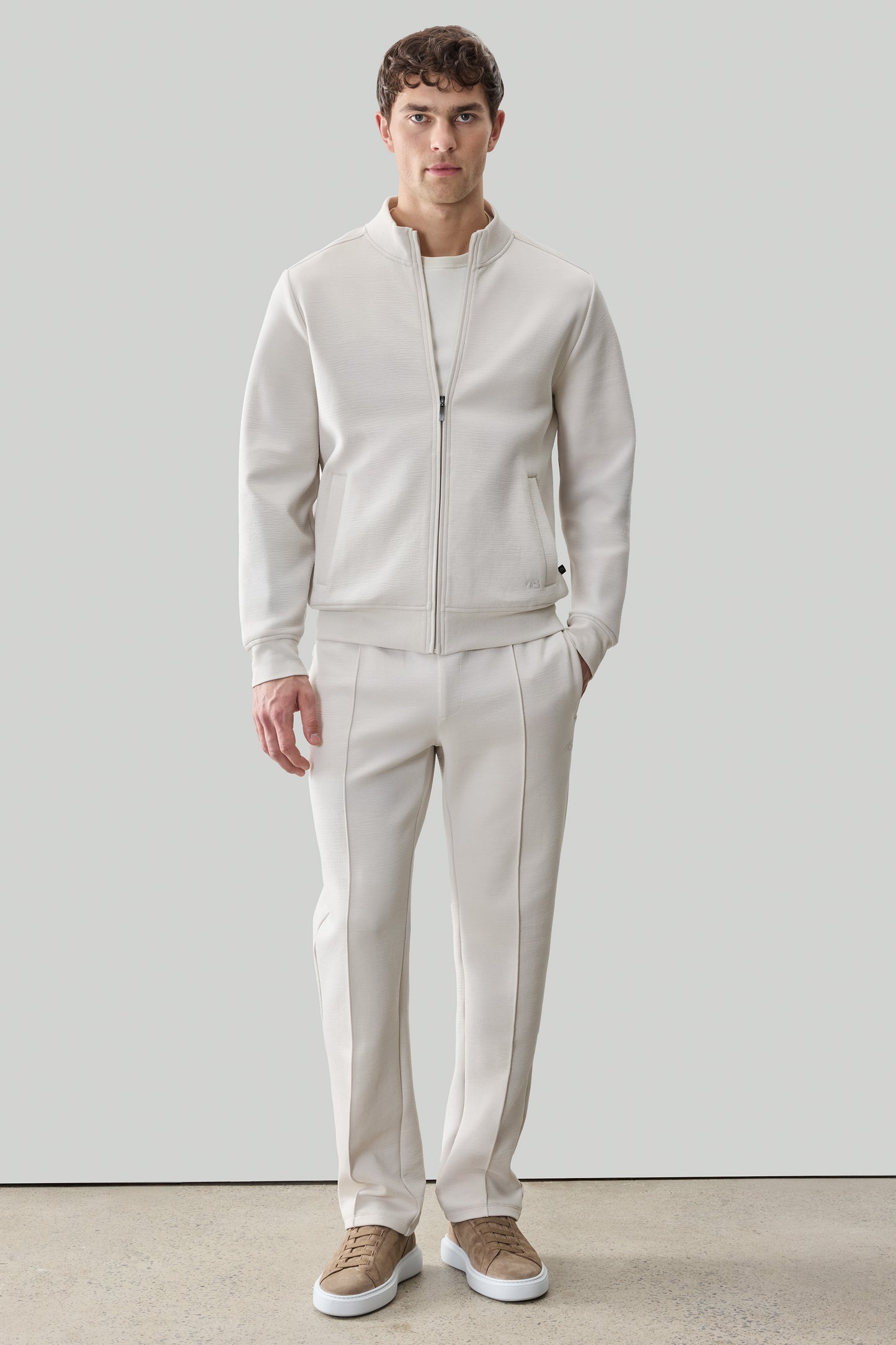 Man wearing cream-colored long sleeve full zip jacket and matching pants with tan sneakers.