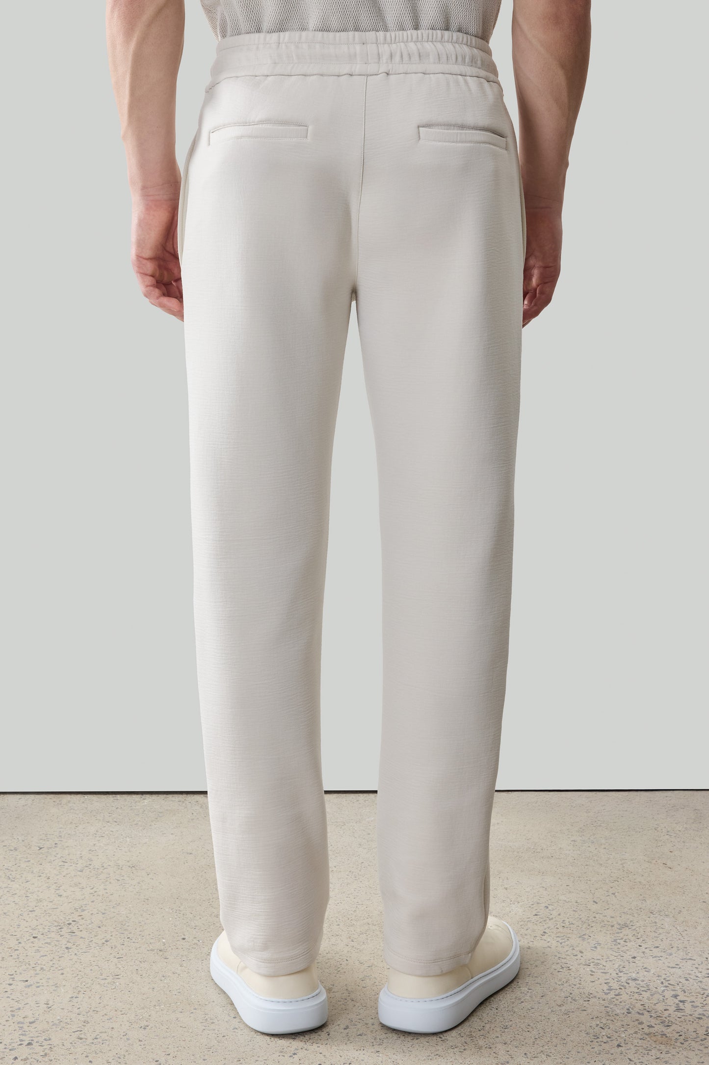 White jogger pants with elastic waistband and back pockets, shown from rear view on model wearing white sneakers.