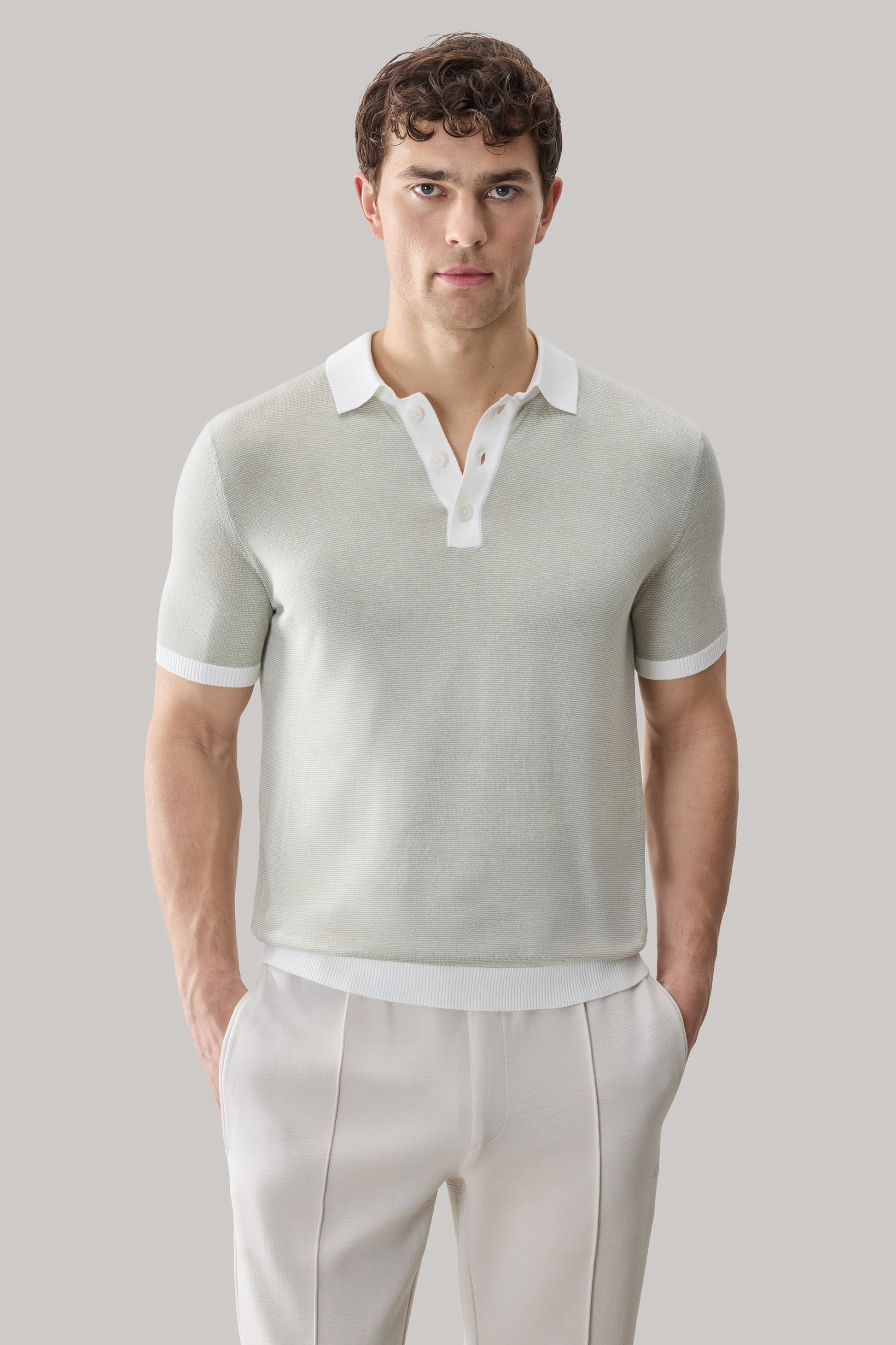 Man wearing sage green short-sleeve sweater polo with white collar and cuff trim over cream pants.