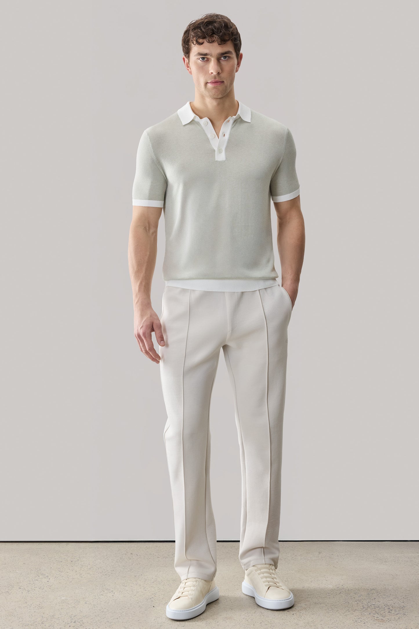 Man wearing light beige short-sleeve sweater polo with white collar and cuffs, paired with cream trousers and white sneakers.