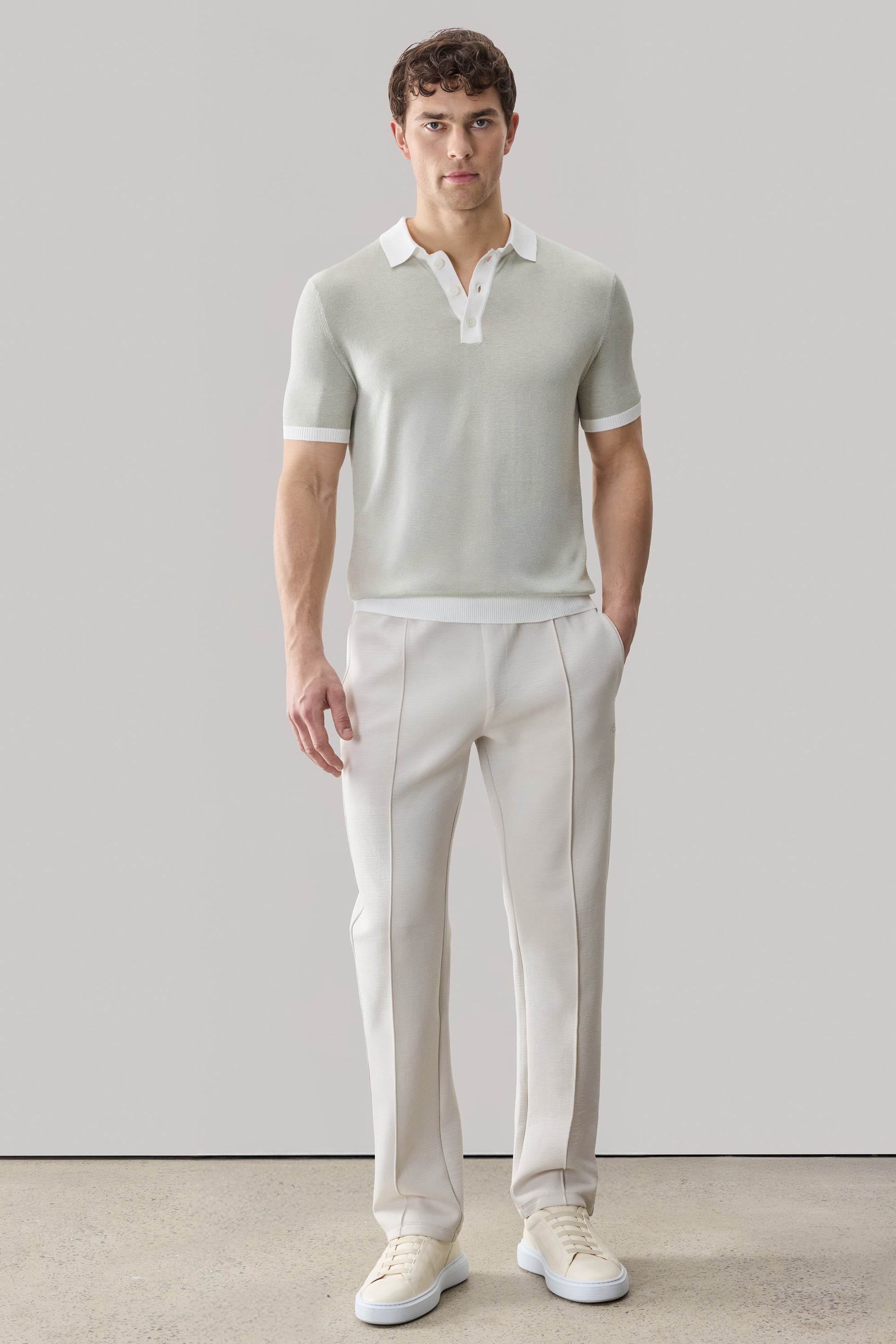 Man wearing light beige short-sleeve sweater polo with white collar and cuffs, paired with cream trousers and white sneakers.