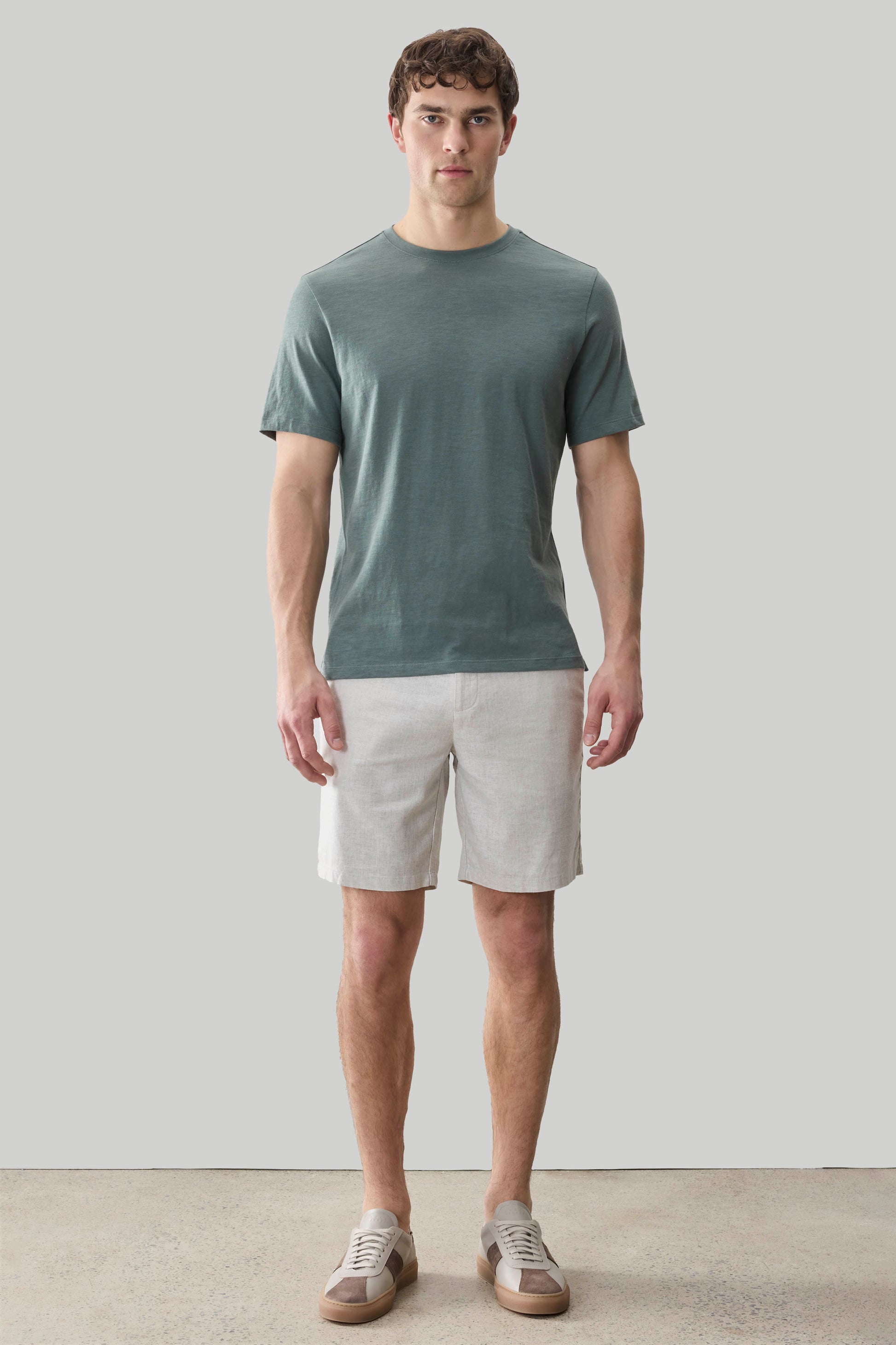 Man wearing a sage green short-sleeve t-shirt with cream shorts and sneakers, standing against a neutral background.