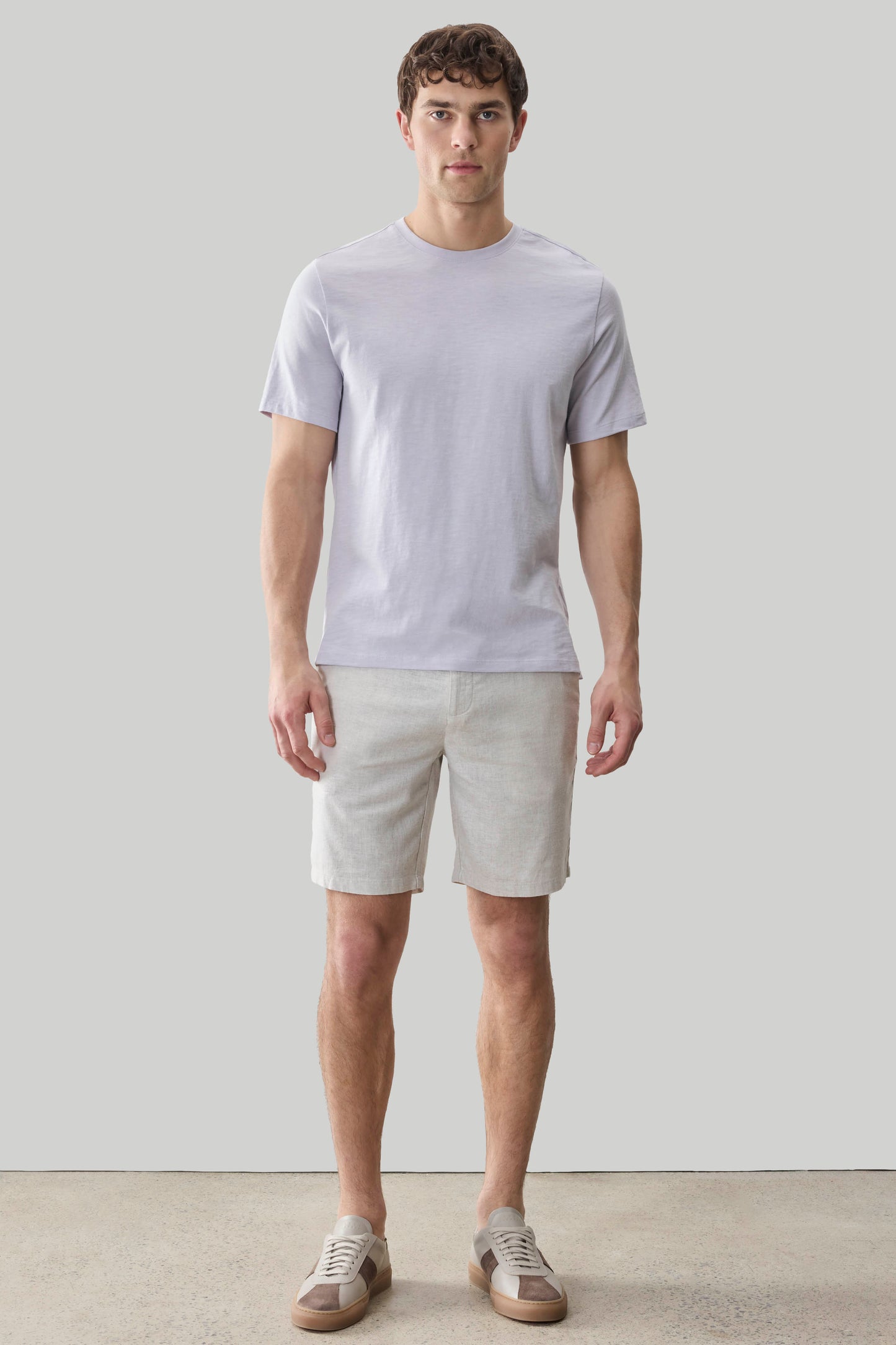 Man wearing light purple short-sleeve crew neck tee with beige shorts and canvas sneakers.