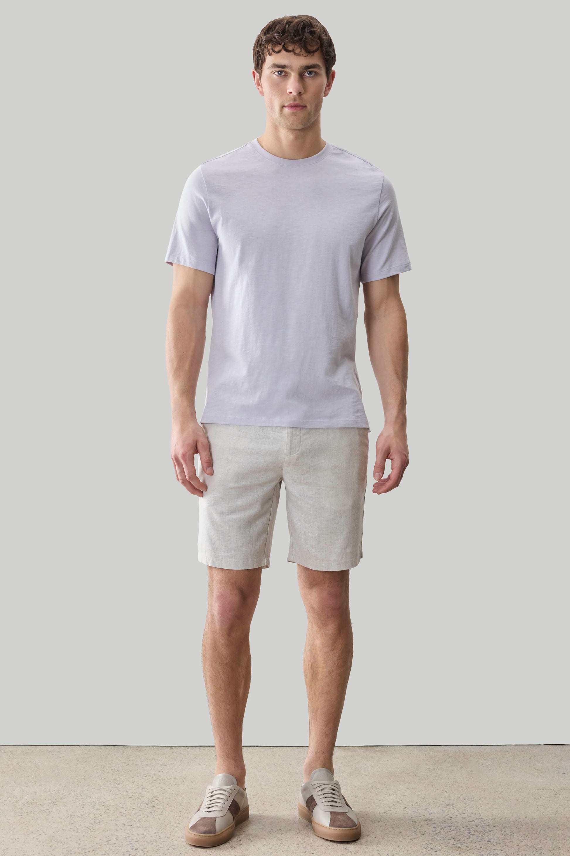 Man wearing light purple short-sleeve crew neck tee with beige shorts and canvas sneakers.
