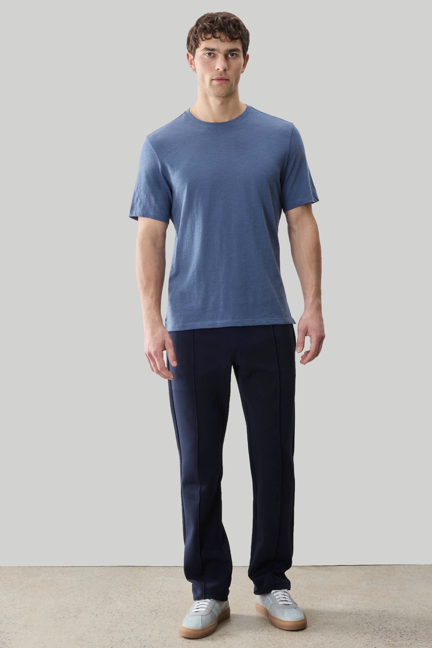 Man wearing a blue short-sleeve crew neck tee and dark navy pants with white sneakers.