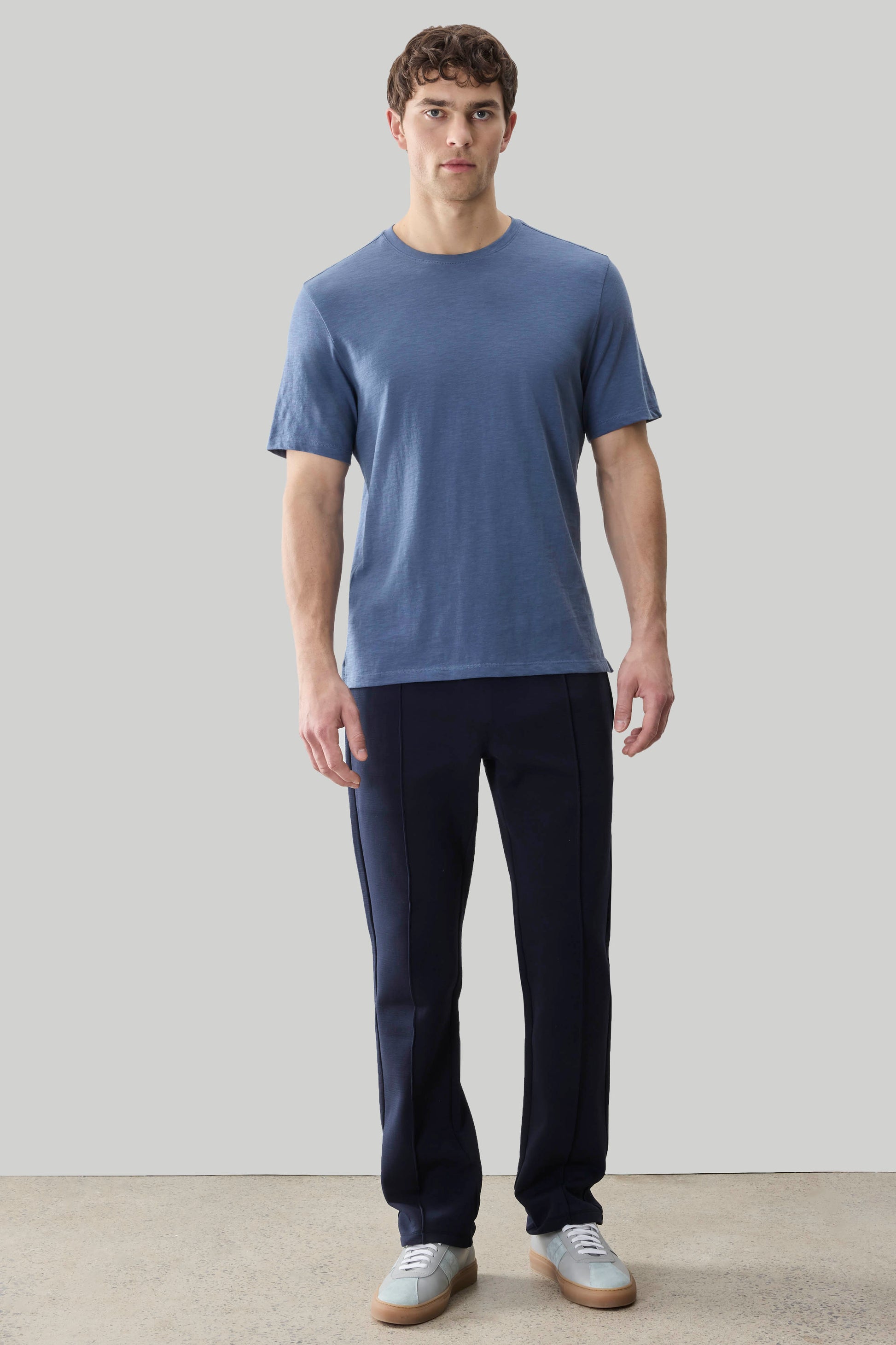 Man wearing a blue short-sleeve crew neck tee and dark navy pants with white sneakers.