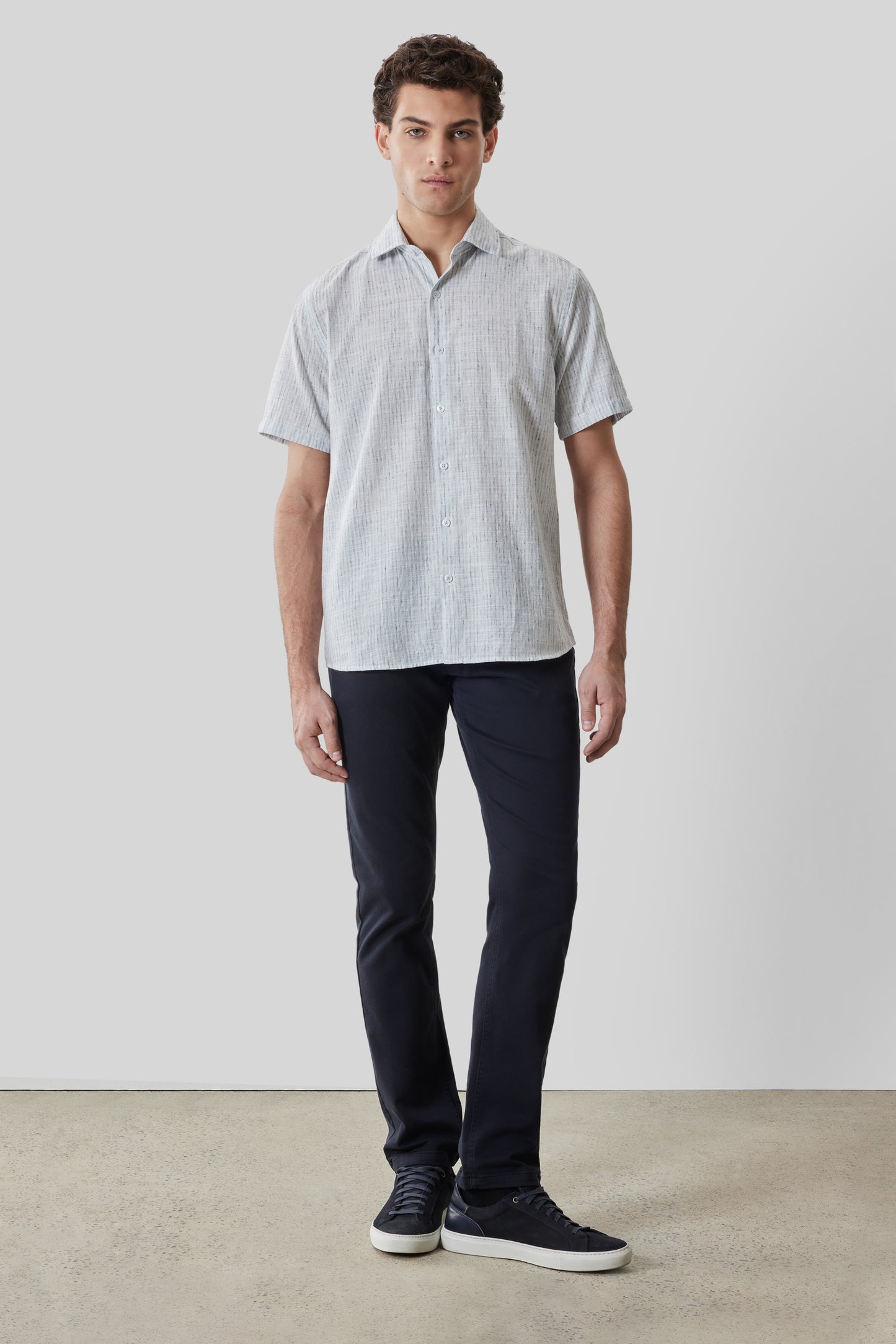 Man wearing light gray striped short-sleeve button-up shirt with dark navy pants and black sneakers.