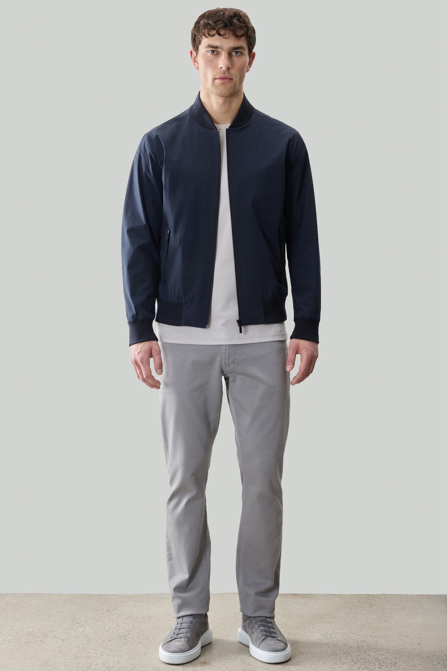 Man wearing navy zip-up jacket with ribbed cuffs over white shirt, gray pants, and gray sneakers.