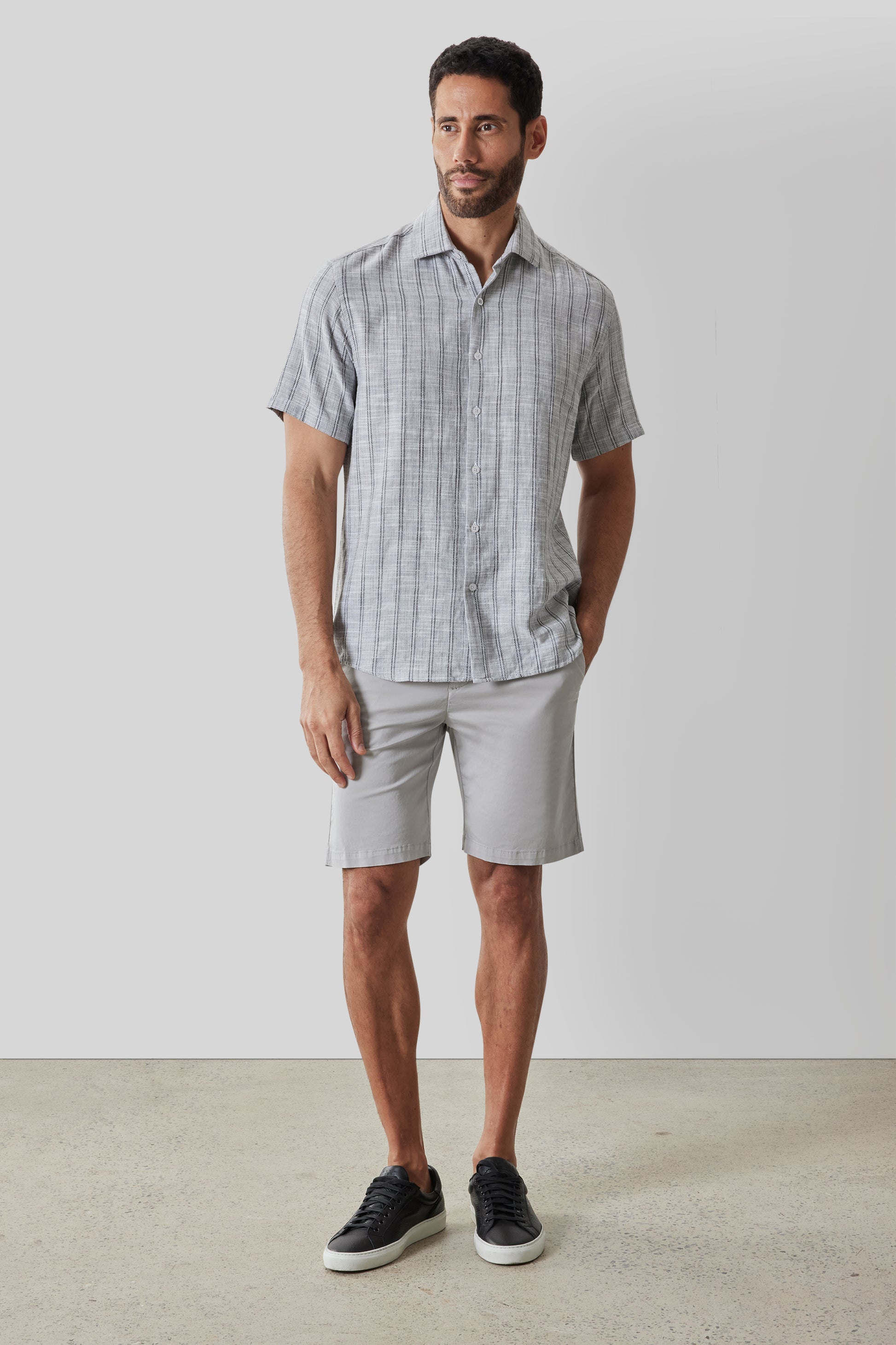 Man wearing gray plaid short-sleeve button-up shirt with light gray shorts and black sneakers.