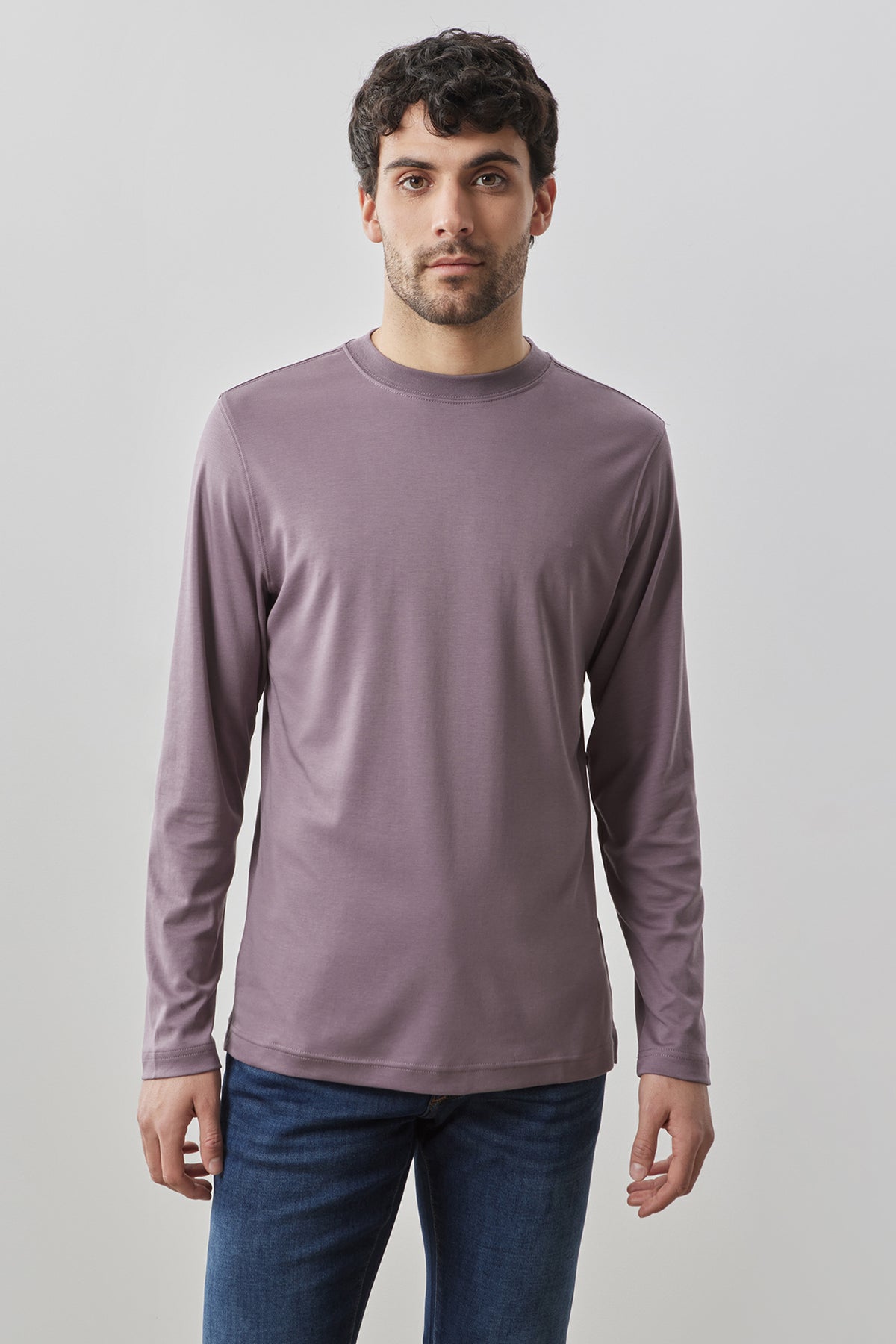 Man wearing mauve long-sleeve crew neck shirt with dark blue jeans against neutral background.
