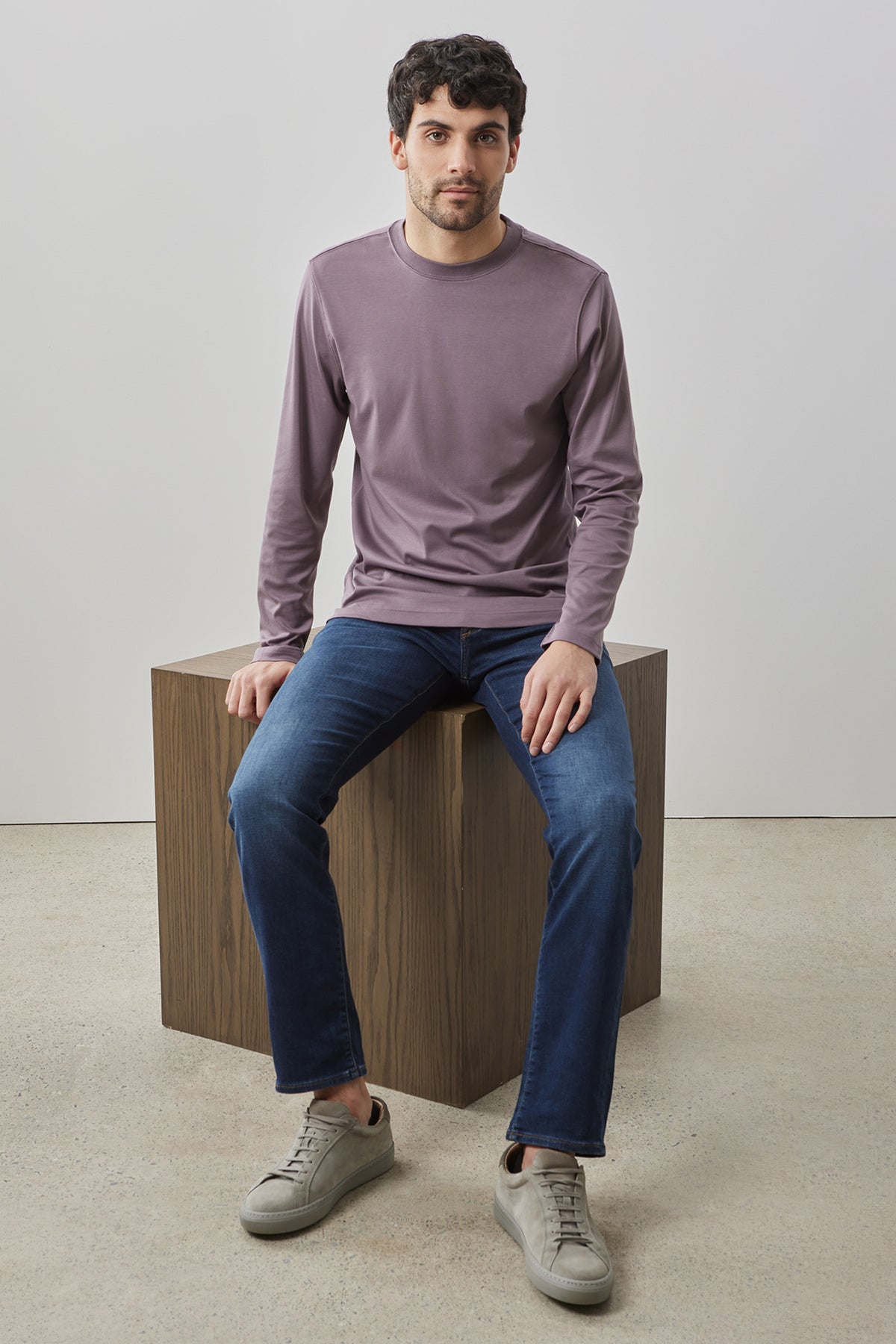 Man wearing mauve long-sleeve crew neck sweatshirt with dark blue jeans and gray sneakers, seated on wooden block.