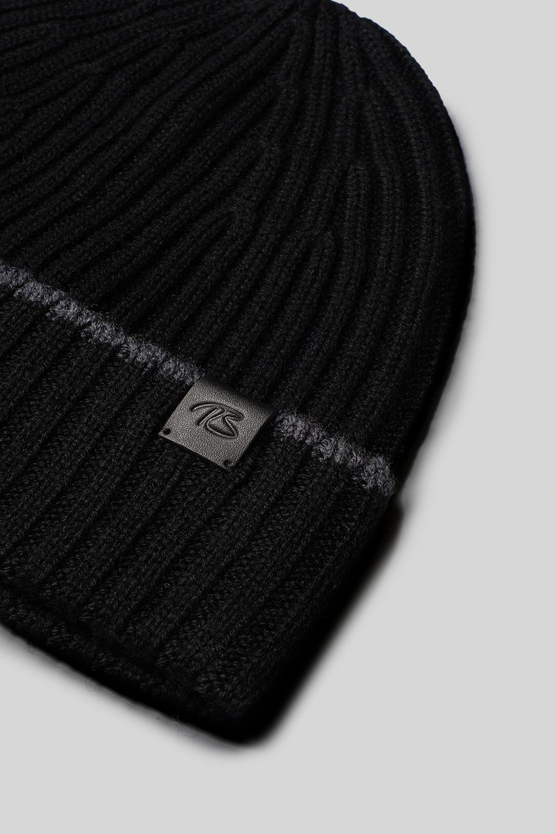 Black ribbed knit hat with metal brand logo patch on cuff.