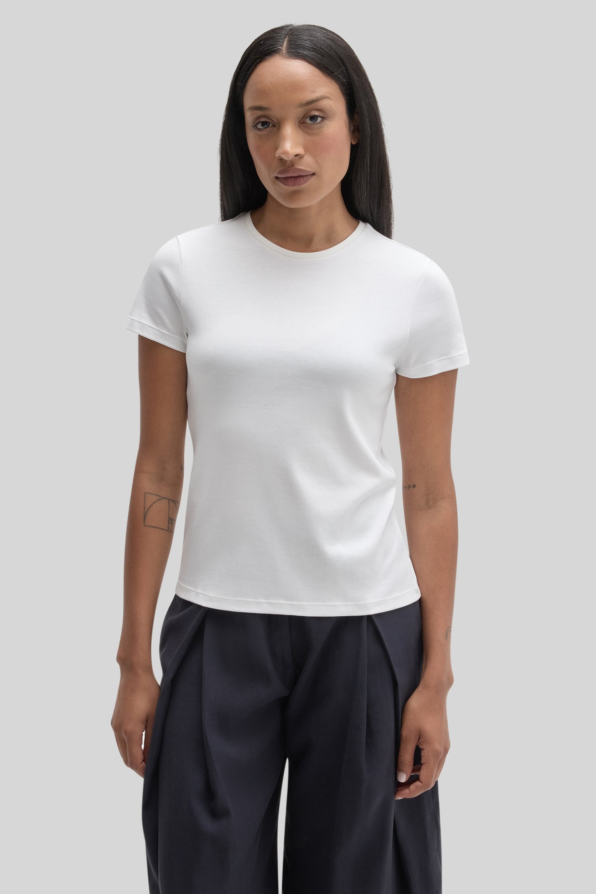 White crew neck short-sleeve tee worn with black pants against a neutral background.