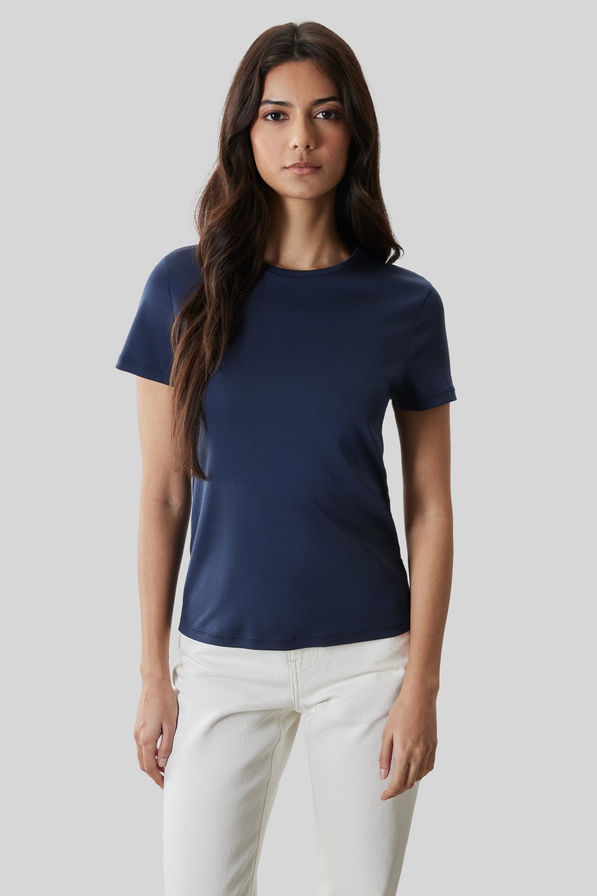 Woman wearing a navy blue crewneck t-shirt with short sleeves and white pants against a neutral background.