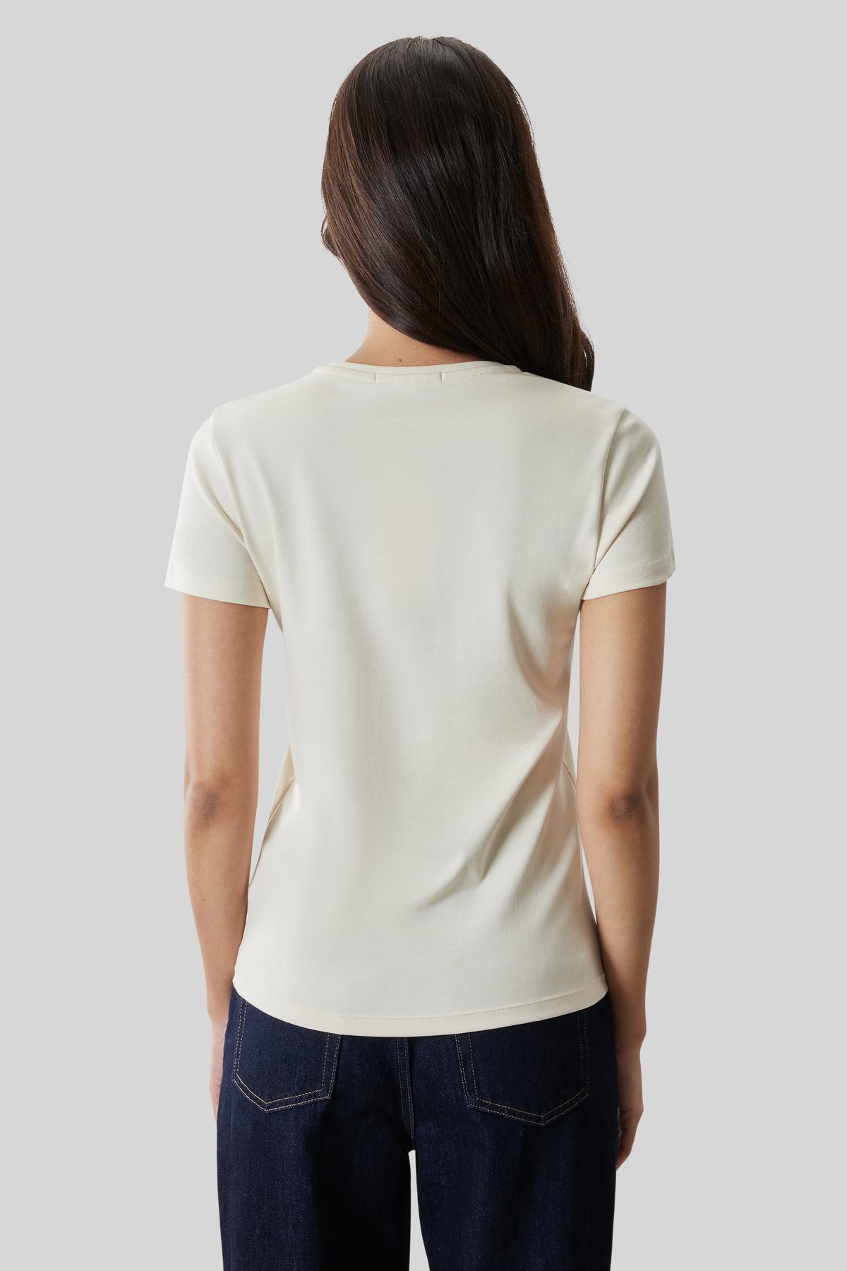 Woman wearing cream short-sleeve t-shirt and dark denim jeans, viewed from back.